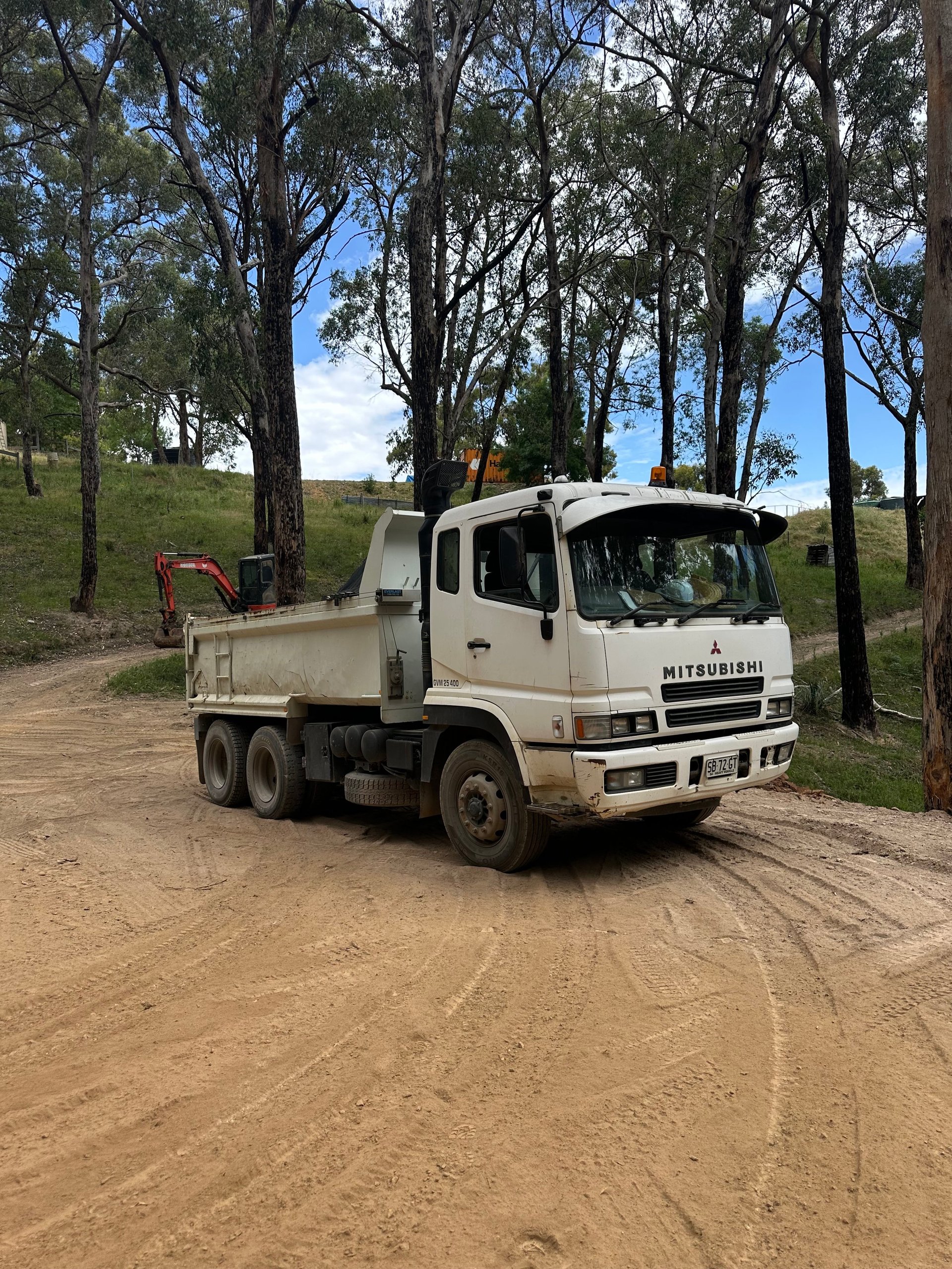 White Mitsubishi dump truck parked on dirt road with tall eucalyptus trees and green hillside in background