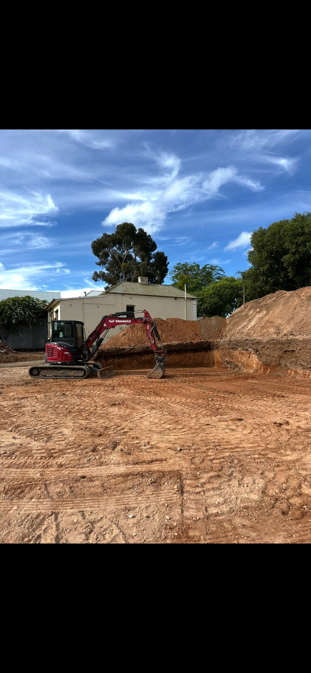 Red excavator loading dirt onto a pile at a construction site under blue sky with white clouds