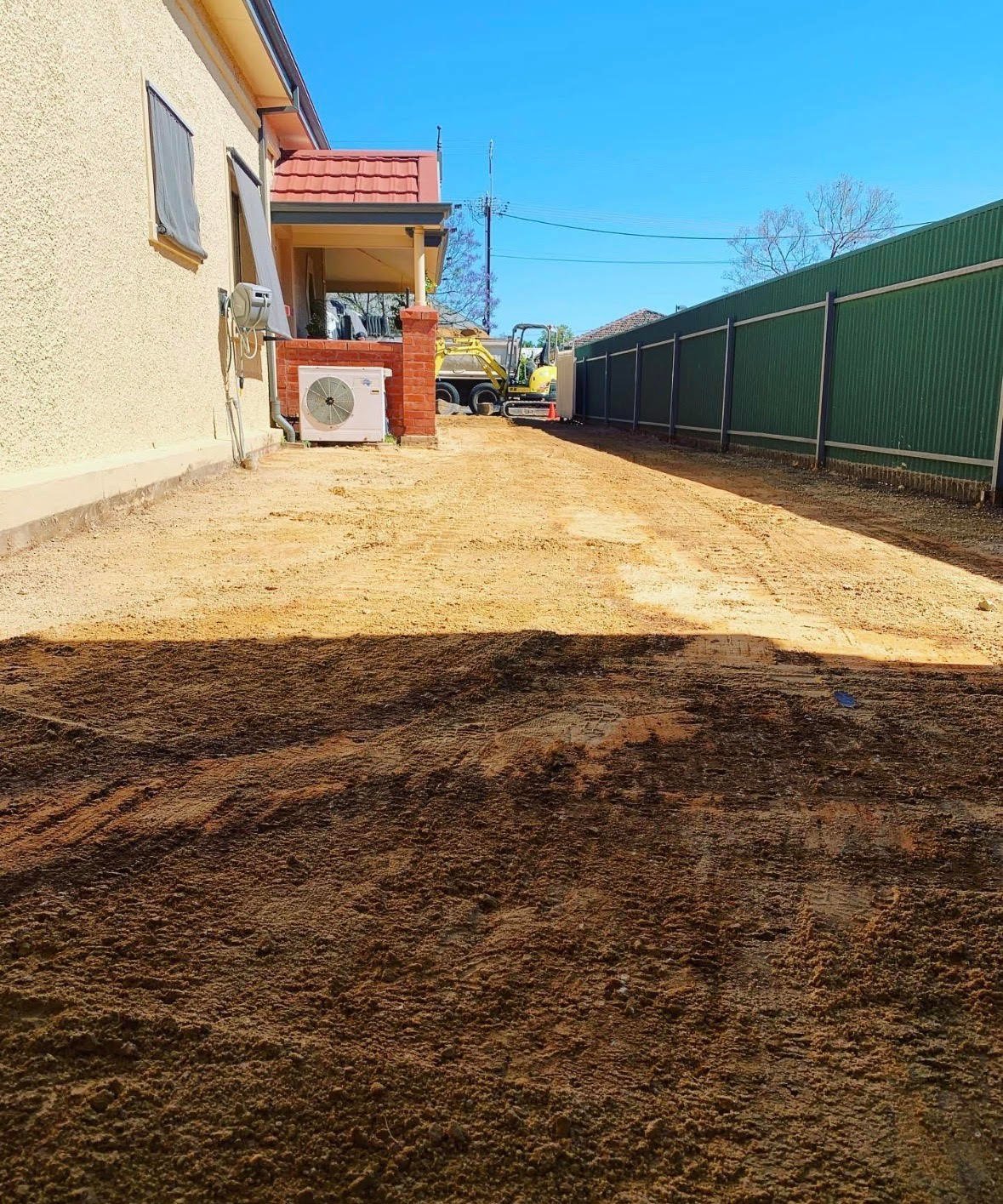 Rural property with cream-colored house, red roof overhang, dirt driveway, green metal fence, and yellow equipment visible on sunny day