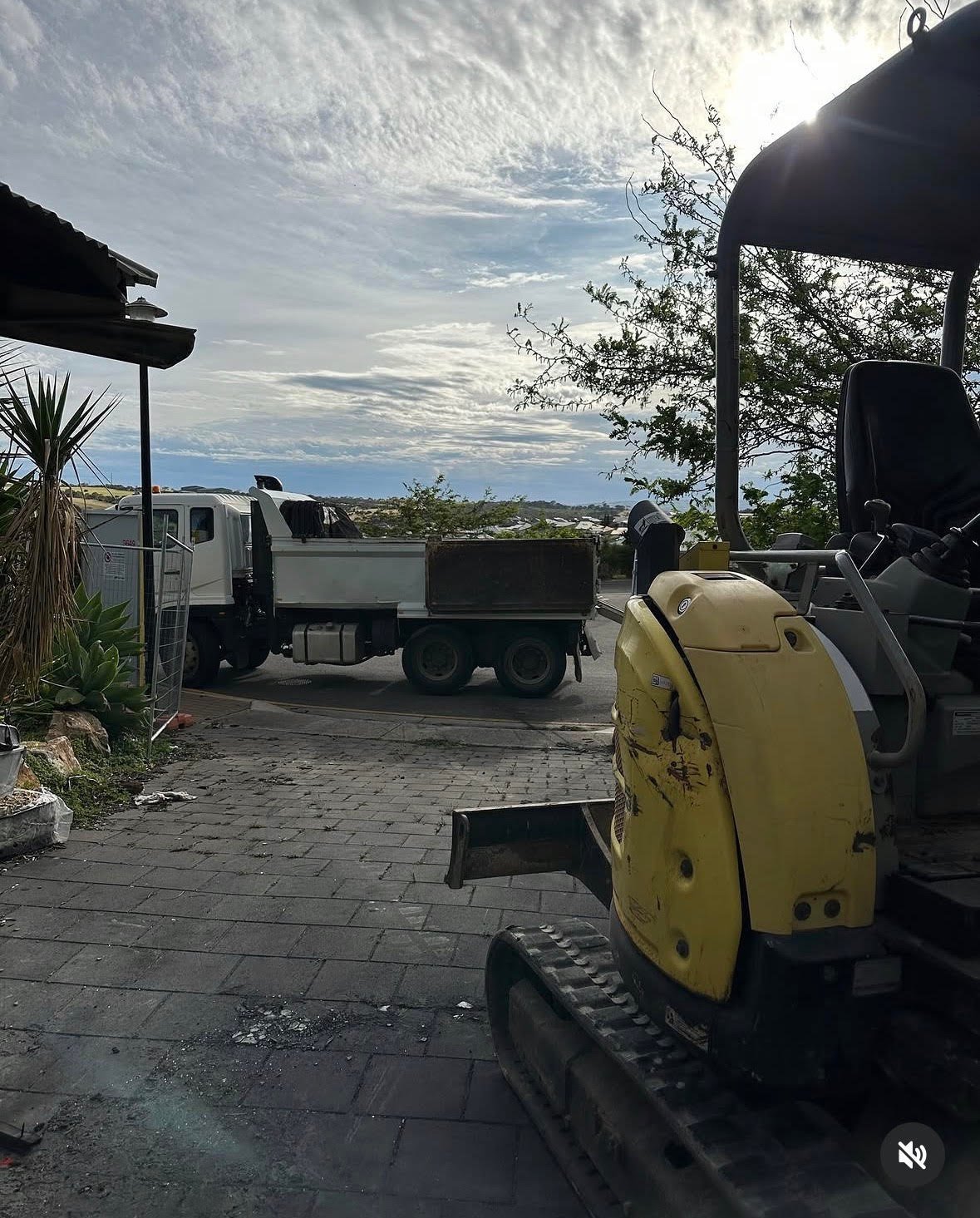 Construction equipment including a yellow excavator and dump truck parked on a paved outdoor area with cloudy sky