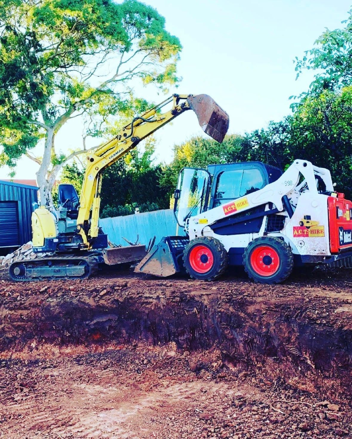 Yellow excavator loading dirt into white and red skid steer loader on construction site with trees and blue shipping container
