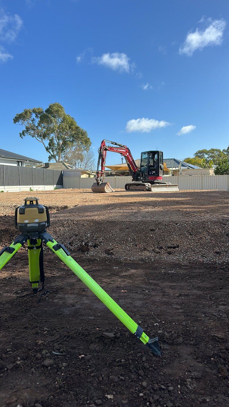 A yellow and green surveying tripod in the foreground with an excavator on a dirt work site under blue sky