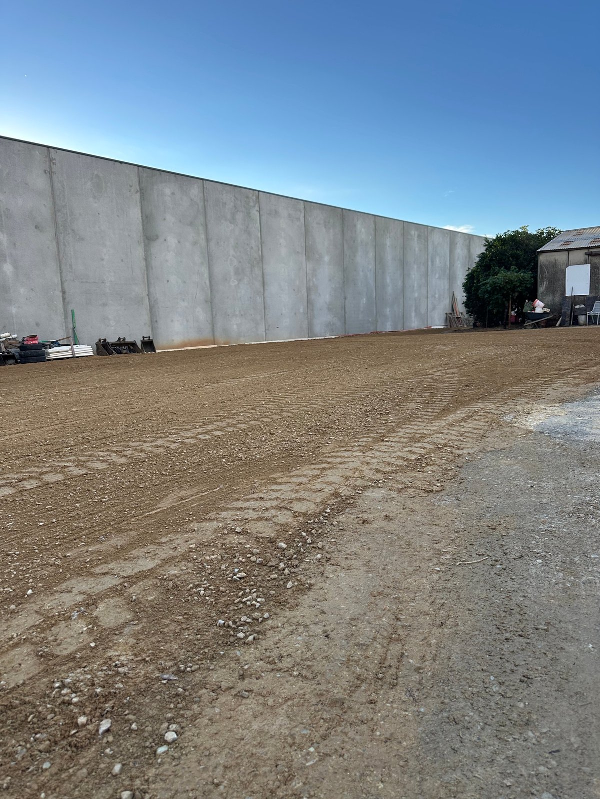 Large concrete wall alongside a dirt parking lot with clear blue sky and industrial buildings in the background