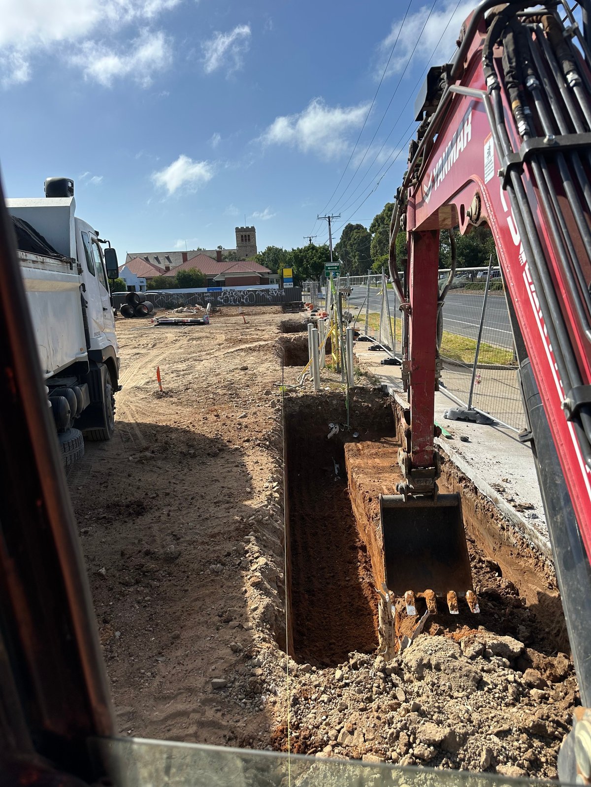 Construction site with red excavator digging in dirt trench, metal fencing, and parked vehicles under blue sky