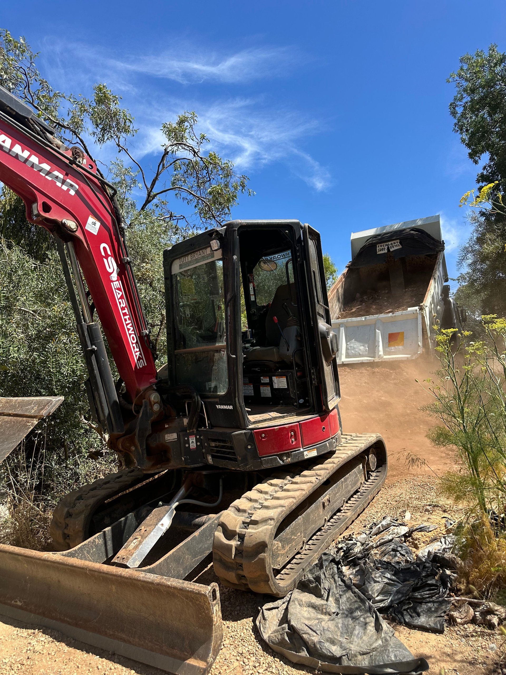 Red and black mini excavator with open cabin door on dirt ground, with dump truck and trees in background