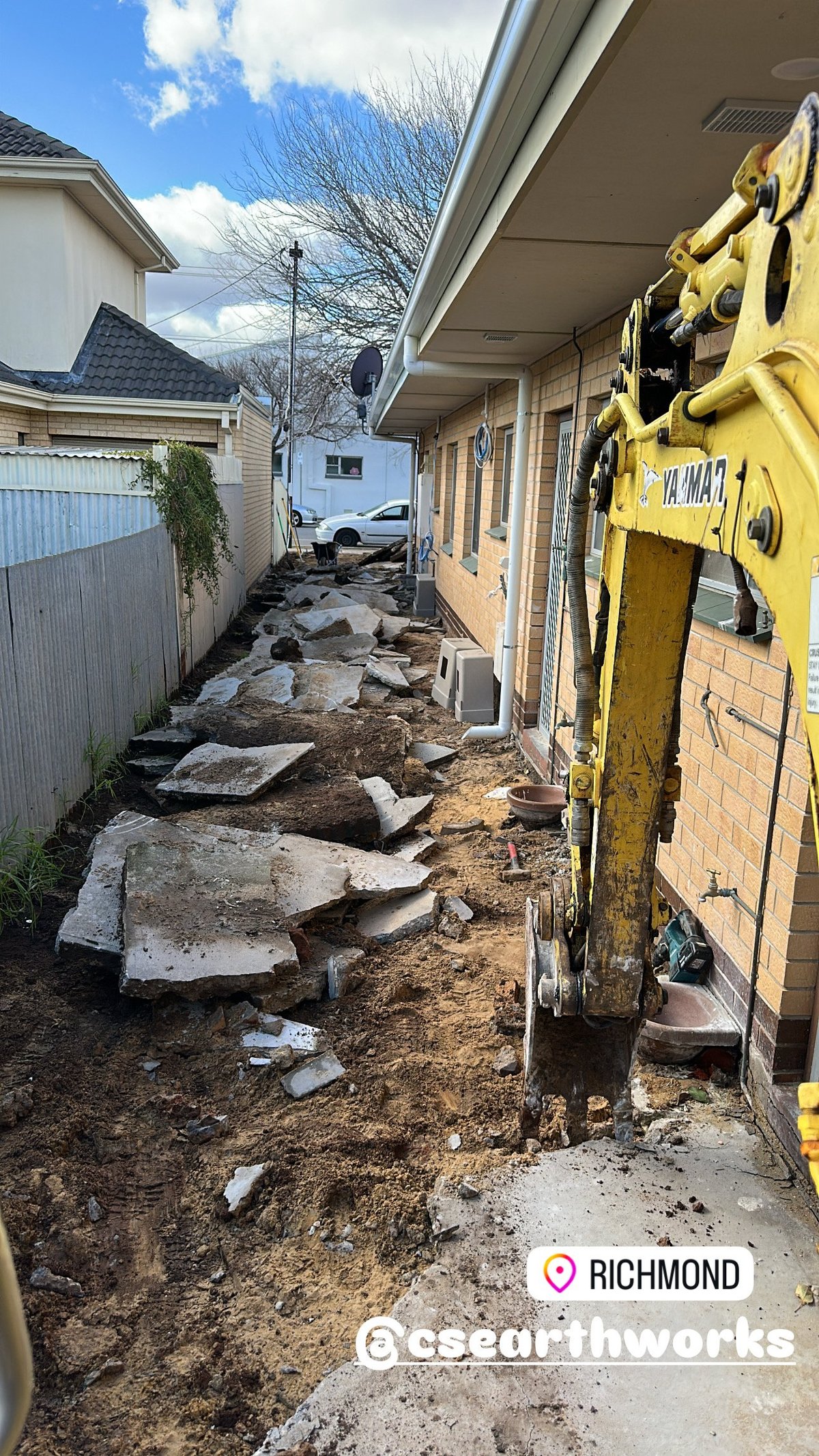 Narrow residential alleyway with exposed dirt, concrete debris, and yellow construction equipment parked along brick buildings