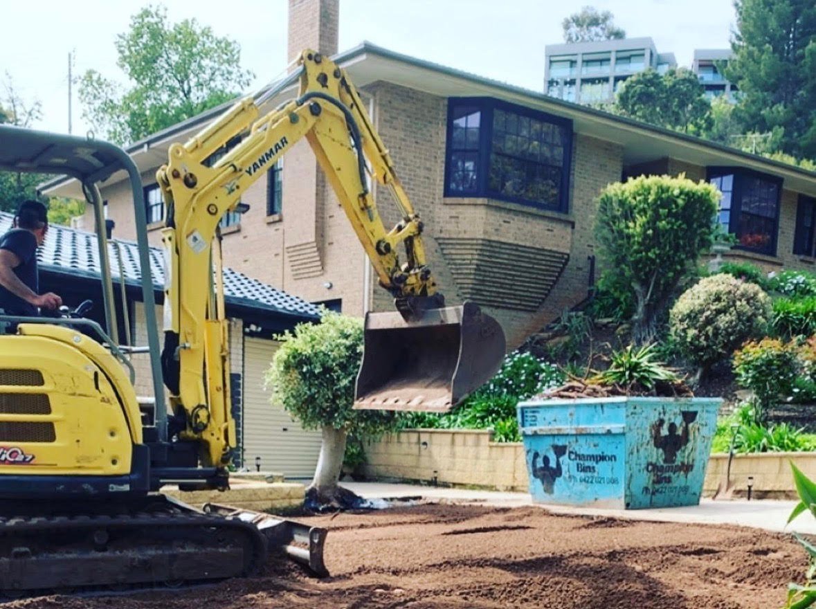 Yellow excavator operating on residential property with brick house and blue container in background