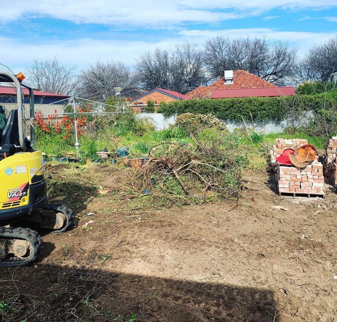 Yellow excavator clearing land with debris pile, brick structure, and residential houses with red roofs in background under blue sky