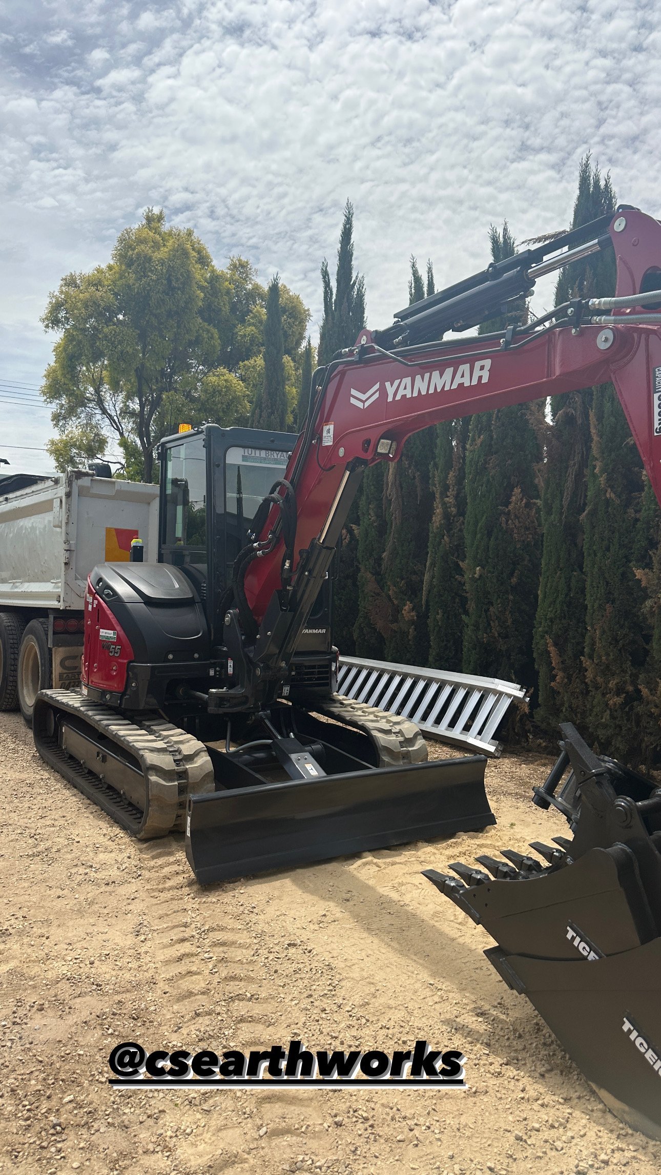 Red Yanmar excavator with front bucket attachment on display with cypress trees and equipment yard in background
